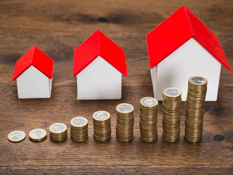 A row of model houses with white walls and red pitched roofs sitting on a wooden table with stacks of coins in front of them. The houses increase in size from left to right, and the stacks of coins increase in size also.