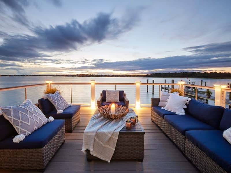 A photograph of a TimberTech deck overlooking a tree-lined lake. The picture is taken at dusk and there are lights in each of the posts of the railing. The guardrail is made from wood-like material and so are the balusters, which are connected with horizontal steel wires that act as a barrier for small pets and children. The decking boards are brown, wood-effect boards with a visible grain. There is a wicker coffee table with a cream colored muslin tablecloth, two metal coffee mugs and a candle burning in an ornate wooden, carved bowl. The tablecloth is pulled back to reveal the front right corner of the coffee table where the coffee mugs are sitting. The surface of the table is made from similar brown wood-like material to the decking. The table is surrounded by wicker furniture with blue and cream-colored cushions.