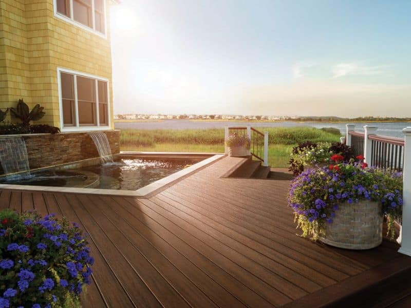 A photograph of Trex decking installed in a home with a view of a nearby river. The decking boards are dark brown and have plan pots arranged on them and steps leading down towards the river. There is a pool with two artificial waterfalls flowing into it sunk into the deck. The pool is right next to the house, which is built from yellow sandstone and has large windows with white frames. The railings around the deck have thin black vertical balusters, white posts, and brown, wood-like guardrails.