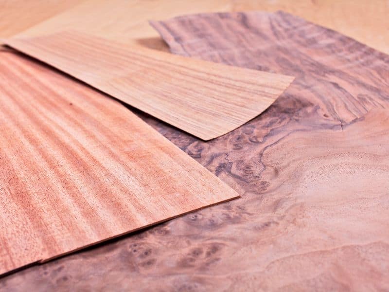 Sheets of brown veneer wood are placed on top of each other. The base sheet appears to be walnut, judging from the grain, and the ones on top could be pine.