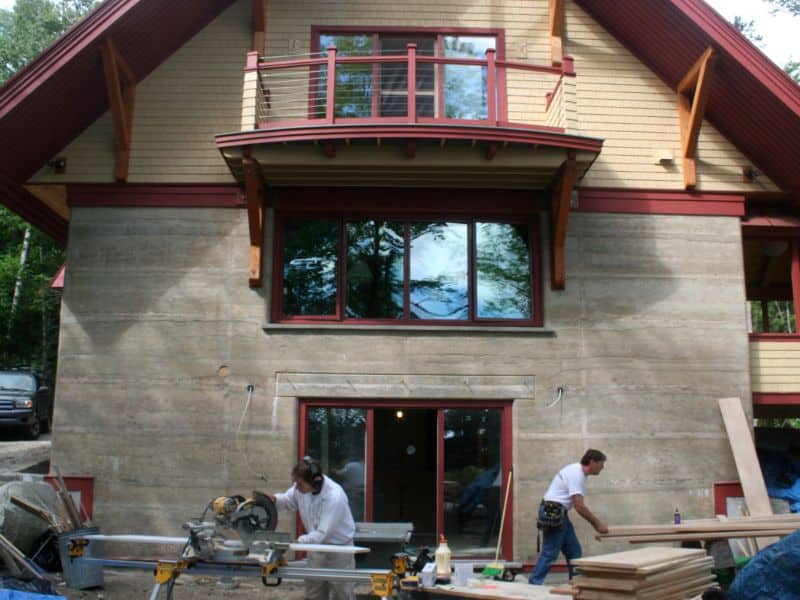 A rammed earth house under construction. The house is three stories high and has red woodwork on the outside. There are two workers outside at ground level cutting and sorting timbers.