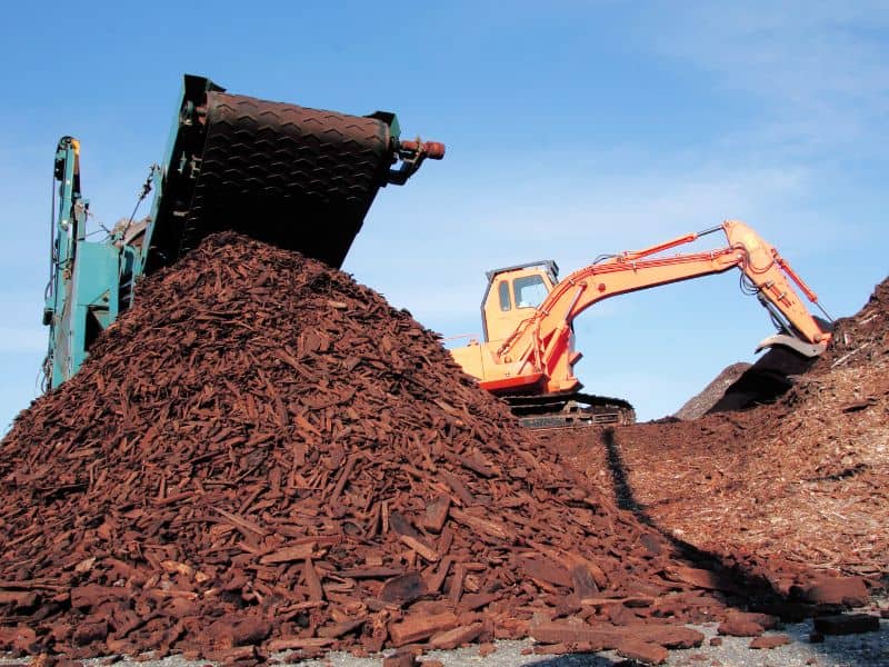 A large pile of scrap wood in a pile underneath a conveyor belt. An orange excavator is moving the wood around.