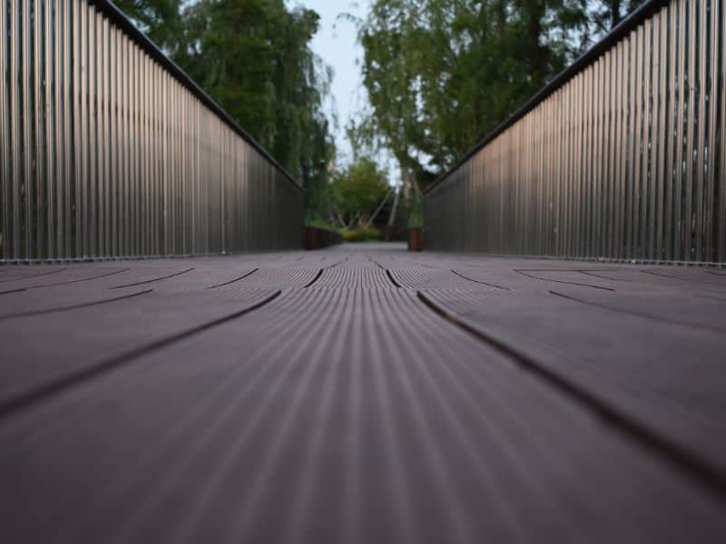 A photograph of warped composite decking taken from a very low angle looking along the length of the boards to the undulations are clearly visible. The boards have grooves running along their lengths. The boards are dark brown in color and look like wood. Either side of the decking walkway is a railing made from wood with light brown balusters and darker brown guardrails. There are leafy green trees either side of the decking.