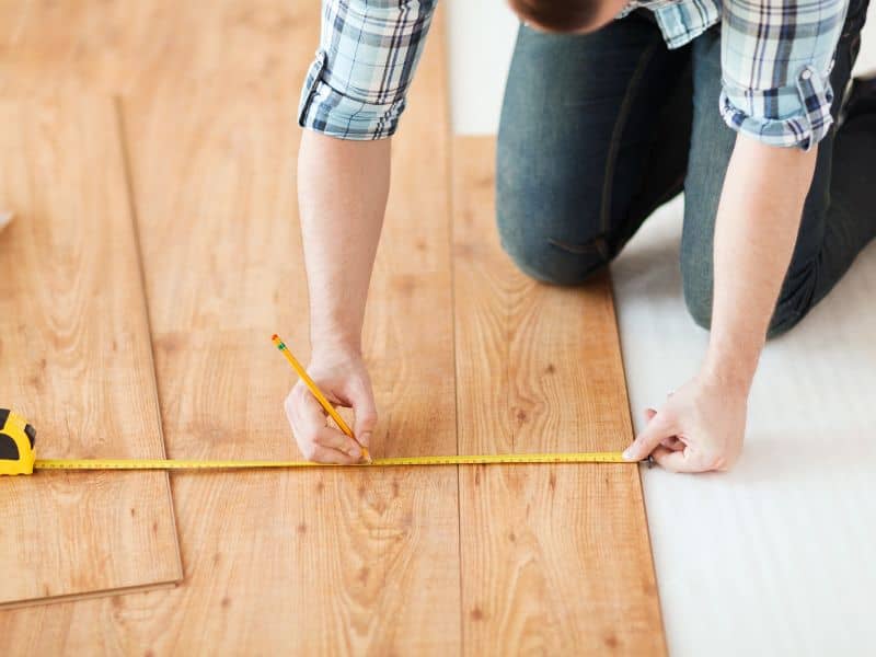 A man kneeling down on wood flooring measuring the boards with a yellow tape measure and marking them with a pencil.