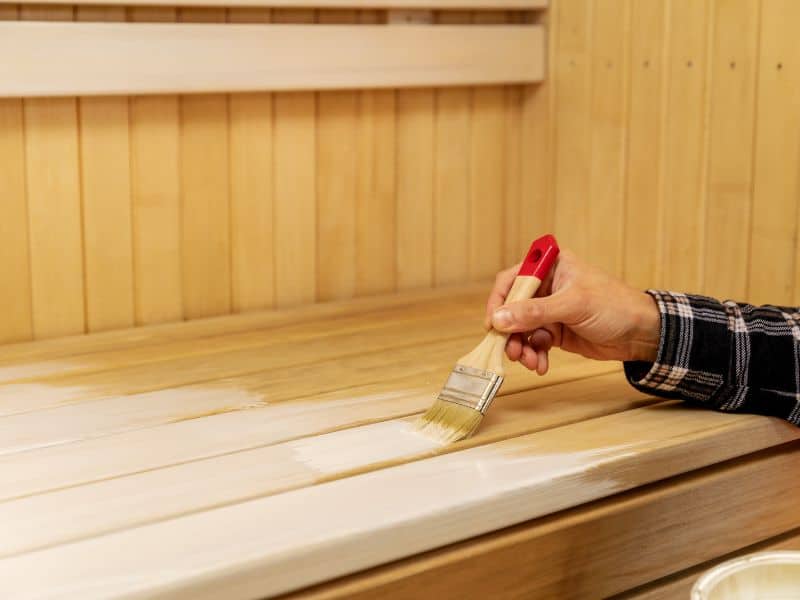 Using a paintbrush, a man applies a wax finish to a wooden bench in a sauna.