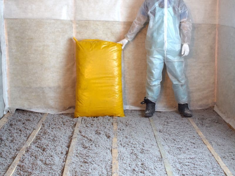 A man wearing a set of see-through overalls standing on floor joists between which cellulose insulation has been placed. There is a large yellow plastic bag full of cellulose standing against the wall, and the man has his hand on top of it.