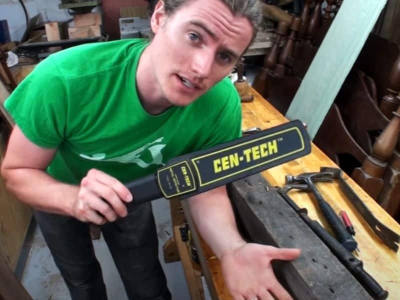 Using a hand-held metal detector, a man is checking reclaimed wood for nails and fasteners. The image is a screenshot from a YouTube video.