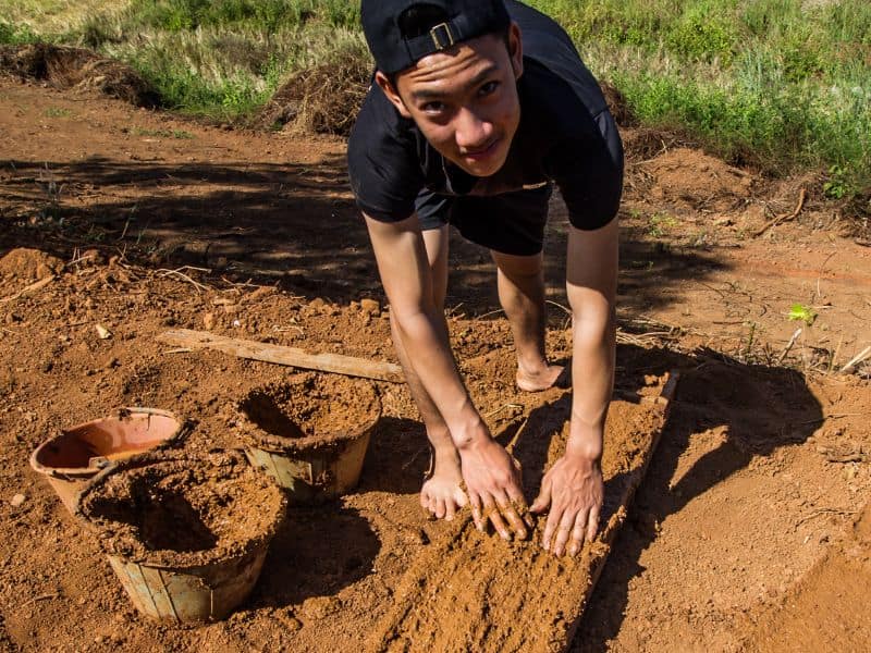 A young man wearing shorts and a T-shirt filling an adobe brick mold with adobe mixture by hand.