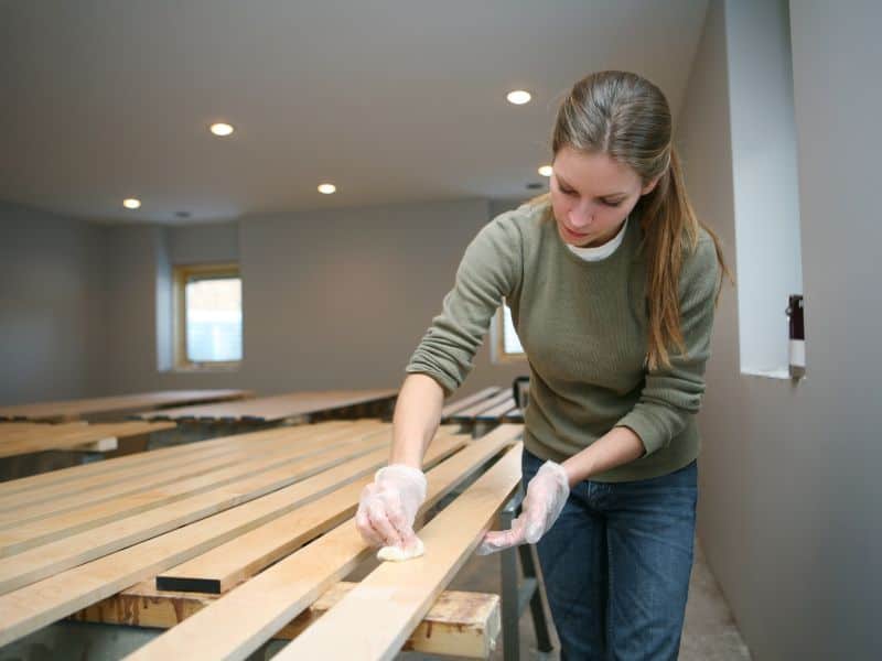 A woman applying gel-based wood stain to planks of wood in a workshop. She is wearing protective plastic gloves and applying the stain with a cotton pad.