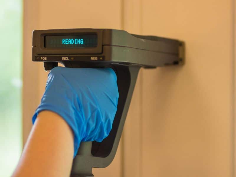 An electronic machine is being held up against a wall to test for the presence of lead in the paint. The machine operator is wearing blue rubber gloves, and the machine display says, "reading."