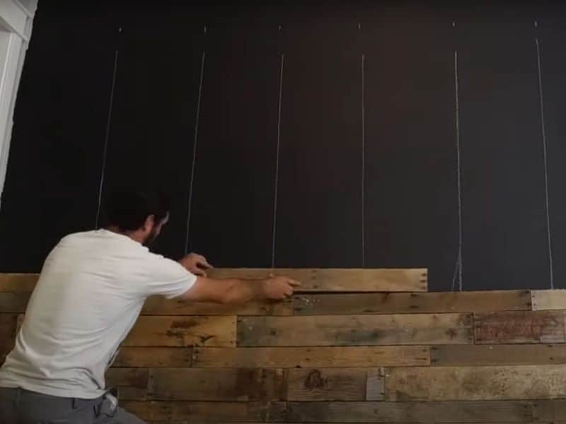 A screenshot of a YouTube video featuring a man placing a reclaimed wood plank on a wall.