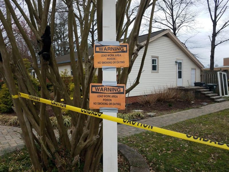 A white, single-story wooden house with a pitched roof. In the foreground are orange warning signs with black writing stating, "Warning! Lead work area. Poison. No smoking or eating." There is also yellow tape with the word "caution" in black letters that has been used to cordon off the area.