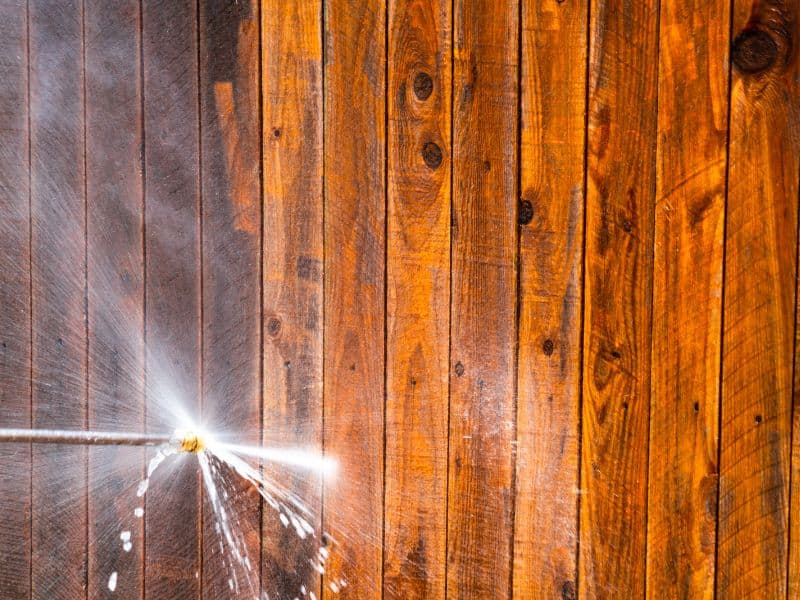 A wood surface comprising multiple planks is washed with a pressure washer. The wood to the right of the pressure wash jet is lighter colored, showing it is cleaner.