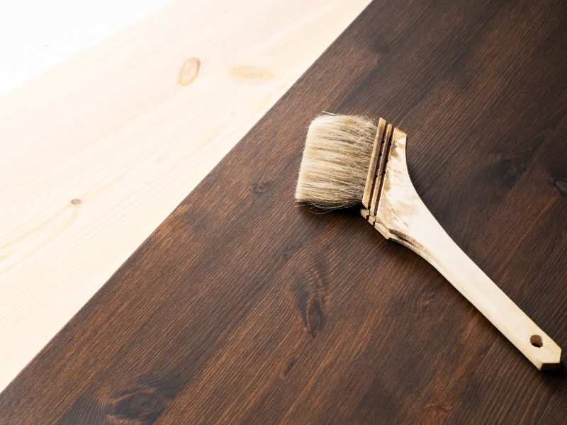 A large paintbrush lies on dark brown stained wood. There is lighter wood next to it to show the difference the staining makes.