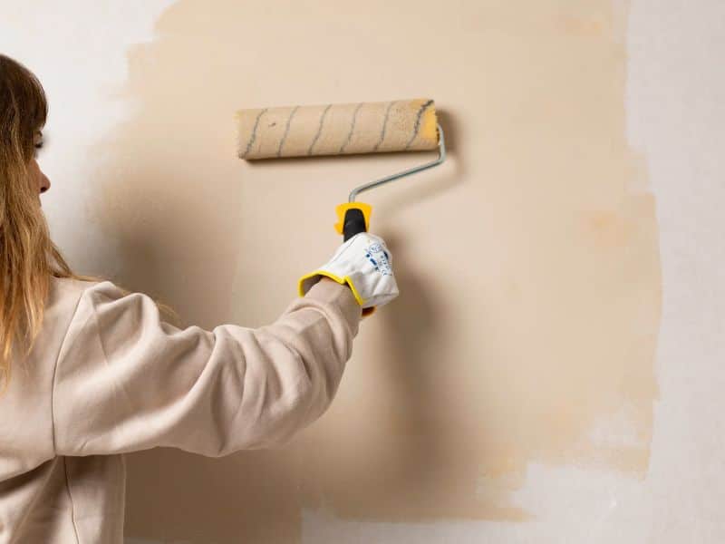 A woman painting a wall light brown with a roller. She is wearing gloves and a light brown sweater.