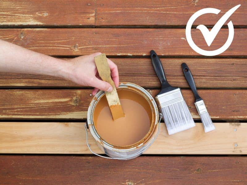 Wooden boards with a tin of wood stain and preservative sitting on top. A man stirs the tin of stain using a piece of wood, and two paintbrushes lie next to the tin.