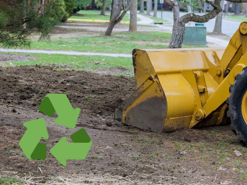 A yellow excavator shovel spreads earth out on the ground. In the bottom left is a recycling icon comprising three circular arrows.