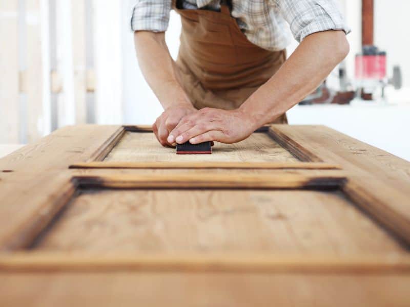 A carpenter sanding down a reclaimed wood door using low-grit sandpaper.