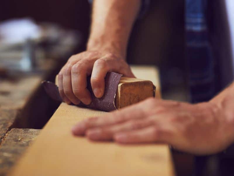 A person is hand sanding a piece of reclaimed wood on a workbench with fine sandpaper and a sanding block.