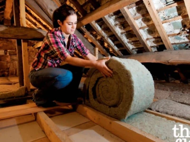 A woman installing a roll of sheep's wool insulation between ceiling joists in a roof space.