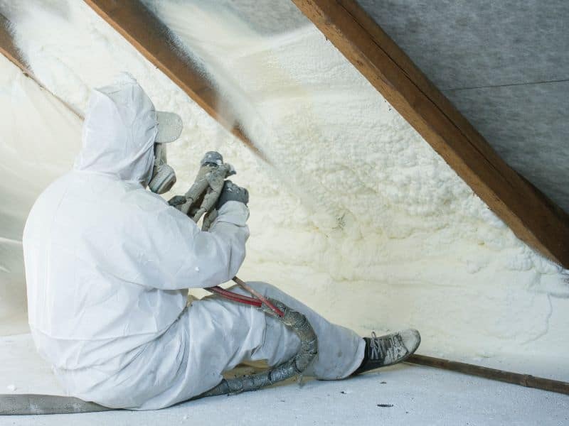 A worker wearing a full PPE kit, including overalls and mask, sitting down in an attic spraying foam insulation between the rafters of the roof.