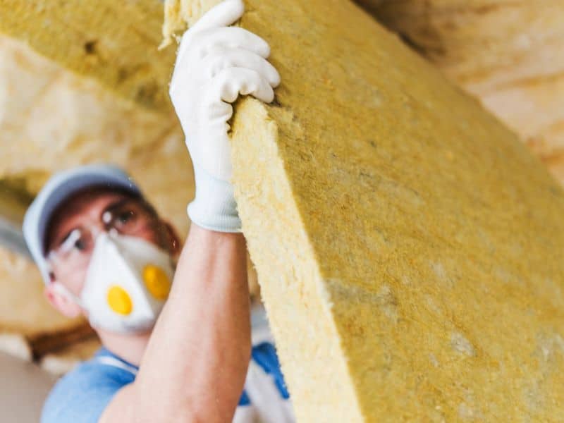 A worker wearing a face mask and protective gloves installing rockwool insulation batts.