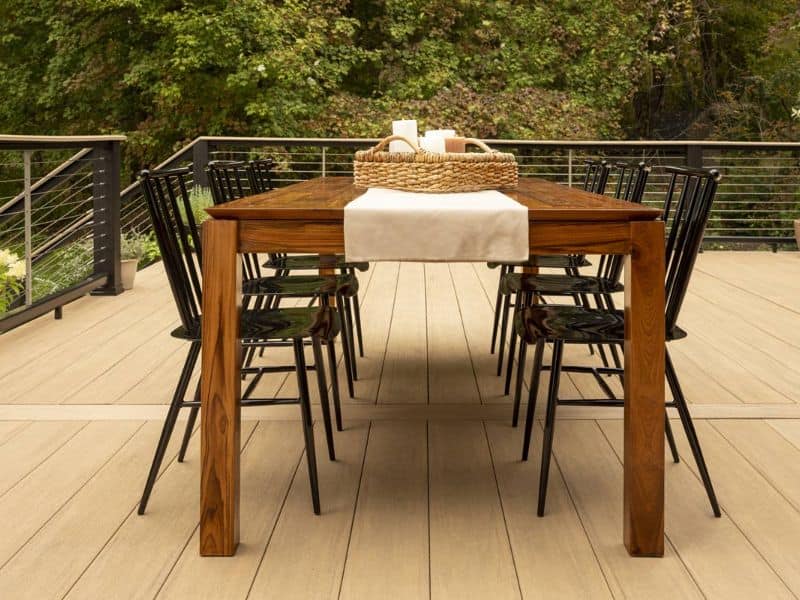 A light brown composite deck on a balcony area overlooking trees. There is a wooden table and chairs with a basket on top of the table.