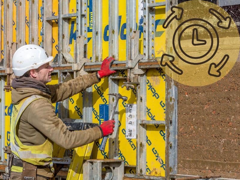 A worker removing metal formwork from a completed rammed earth wall. He wears overalls, a hard hat, and protective gloves and uses a hammer to loosen the fastenings.