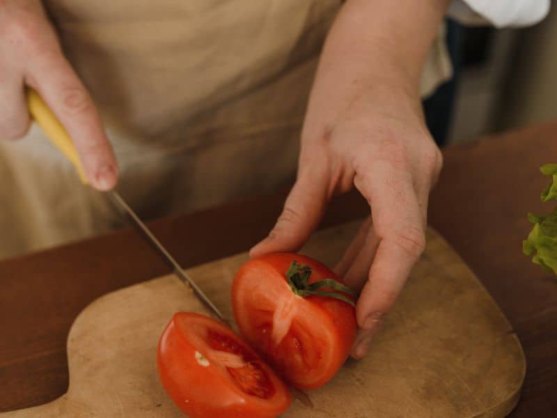 A photograph of a person cutting a tomato on a cutting board on top of a reclaimed wood countertop.