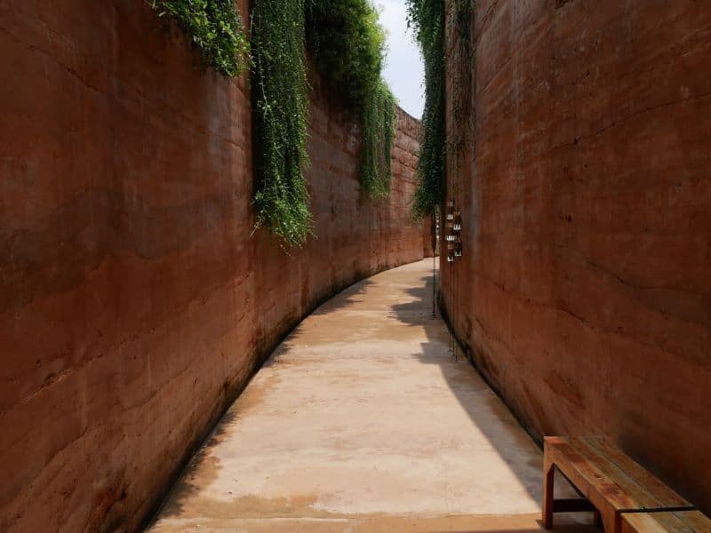 Two tall rammed earth walls made with red-colored earth. The walls are parallel and curve gently to the right making a pleasant walkway that is paved between them. There is vegetation growing down the wall from the top.