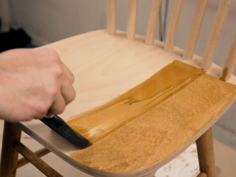 A man's hand with a paintbrush stains the seat of a chair using water-based stain.