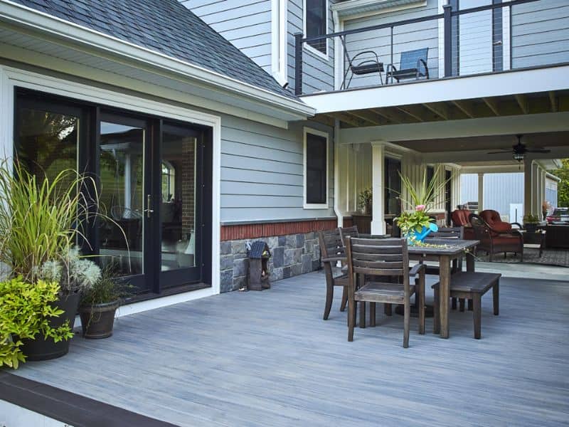 A decked area outside a home with wooden siding and stone cladding. There are tables and chairs on the deck and a balcony above it.