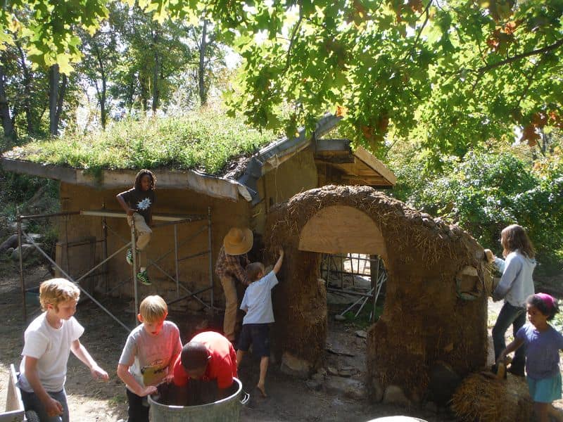 A community builds a cob building with a green roof. Several children are joining in the construction, mixing the cob in a container and applying a course of cob to the walls.