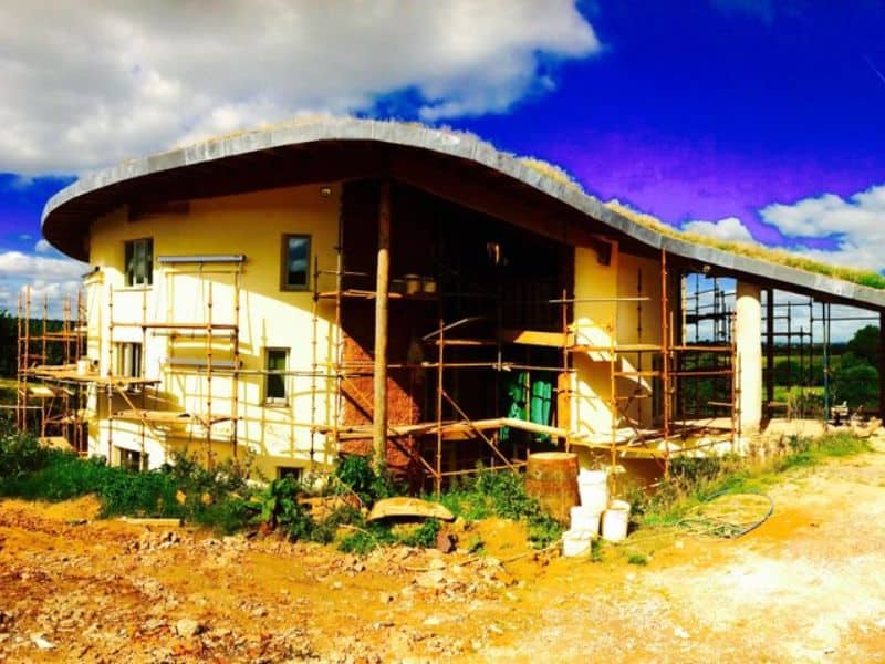 A large cob house, called Dingle Dell, nears completion. The construction site is still active, with scaffolding around the edge of the building and unfinished grounds.