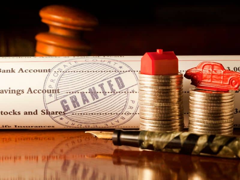 A rolled-up grant document and stacks of coins with a house and a car on top set out on a table. A fountain pen lies on the table in the foreground.