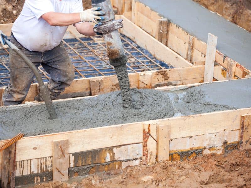 A worker pours a concrete foundation into wooden forms on a building site.