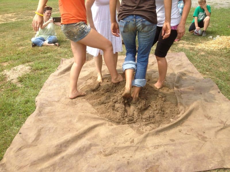 A group of women and children mixing cob with their feet on a tarpaulin laid on the grass.