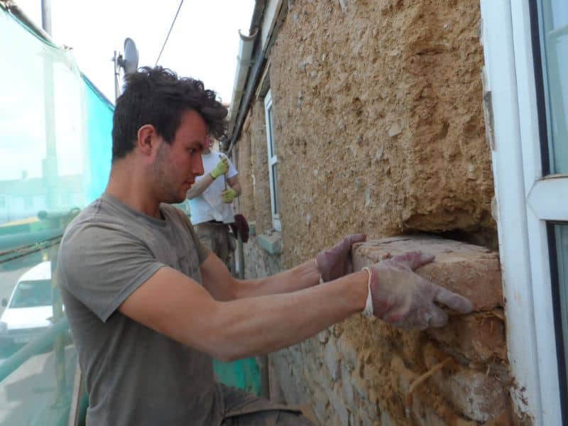 Cob bricks are inserted into the damaged wall as part of a repair. The worker wears protective gloves.