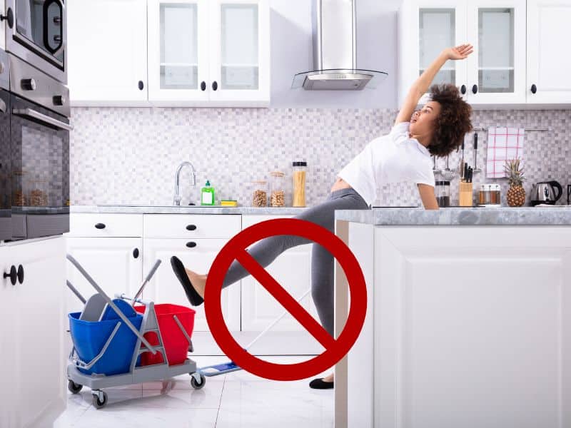 A woman slips on a wet floor in a tiled kitchen. There is a mop and bucket on the floor and the kitchen units are white.