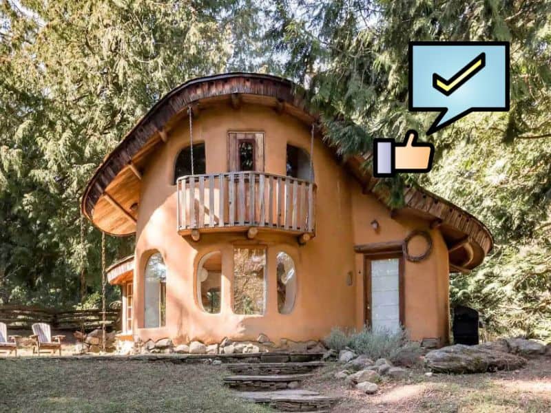 A cob house set in woodland with curved walls and a sweeping curved roof. There is a balcony at the front and a patio area to the left.