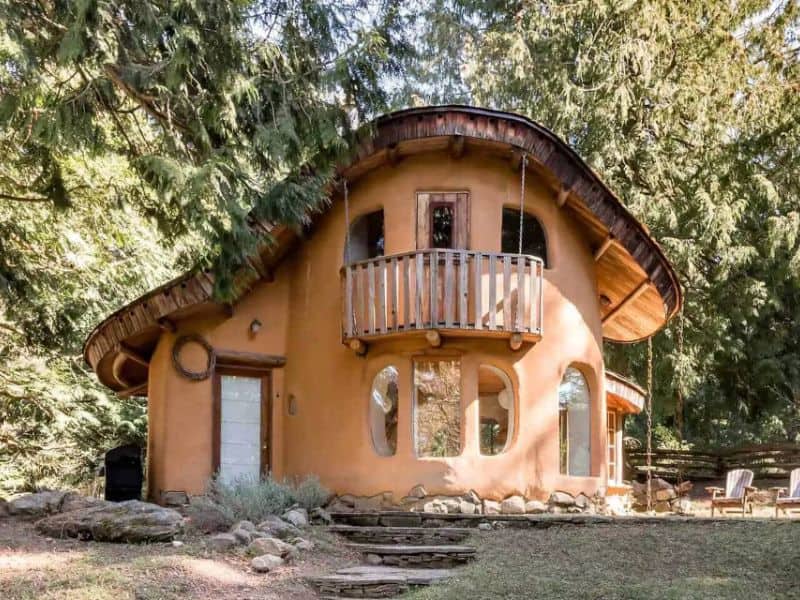 A photograph of a cob house in a forest with a curved façade and irregular-shaped windows. The walls are light orange-brown, and the house has a reclaimed wood balcony.