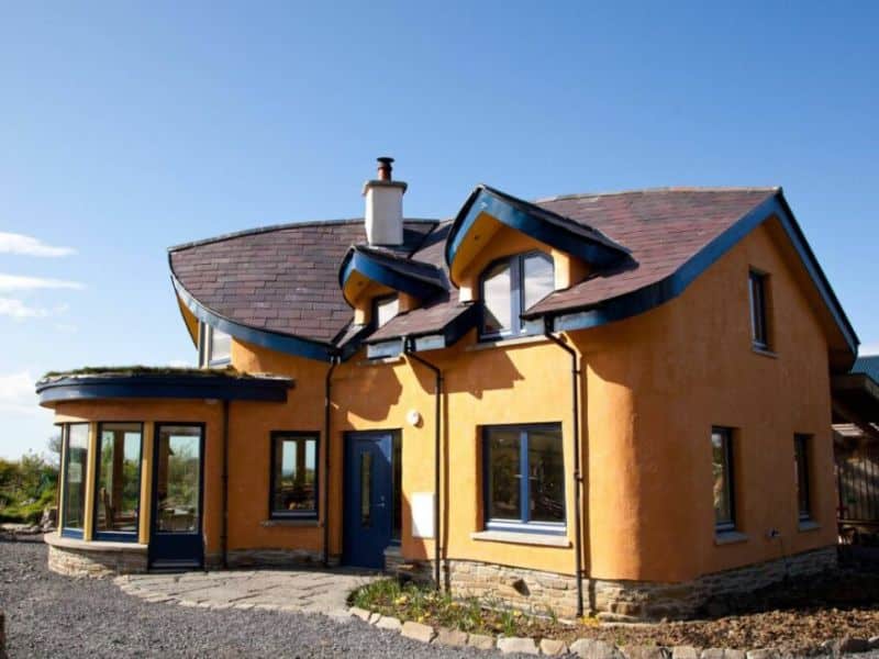 A two-story cob house with orange-rendered walls, large windows, and a curved roof.