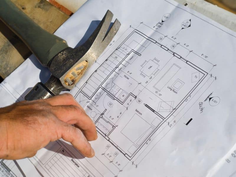 A set of house plans laid out on planks of wood with a hammer holding them down and a builder's hand pointing to our details.