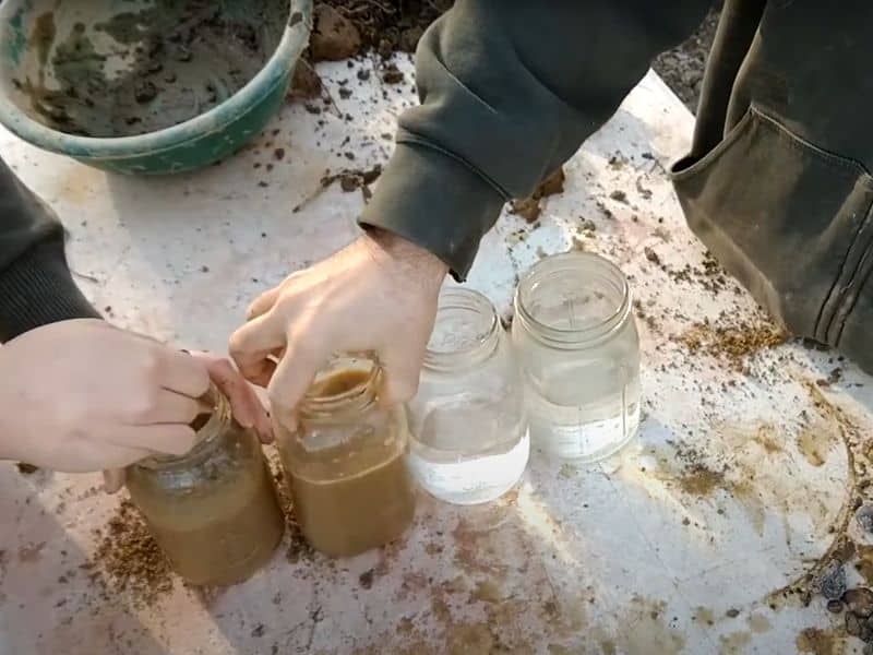 A screenshot of a YouTube video on the Uncle Mud channel showing people working with jars full of mud and water during a soil shake test.