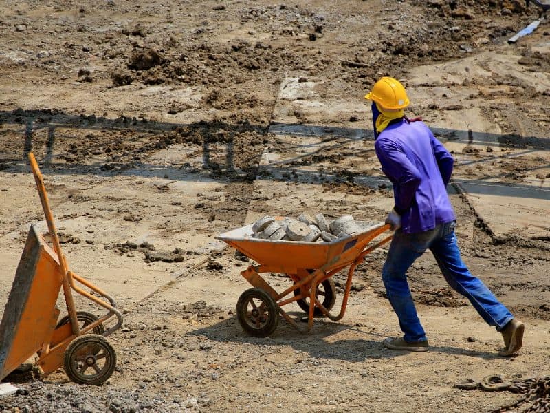 A worker on a conventional construction site is pushing a wheelbarrow full of concrete cores. He wears a yellow hard hat and blue overalls, and blue jeans.