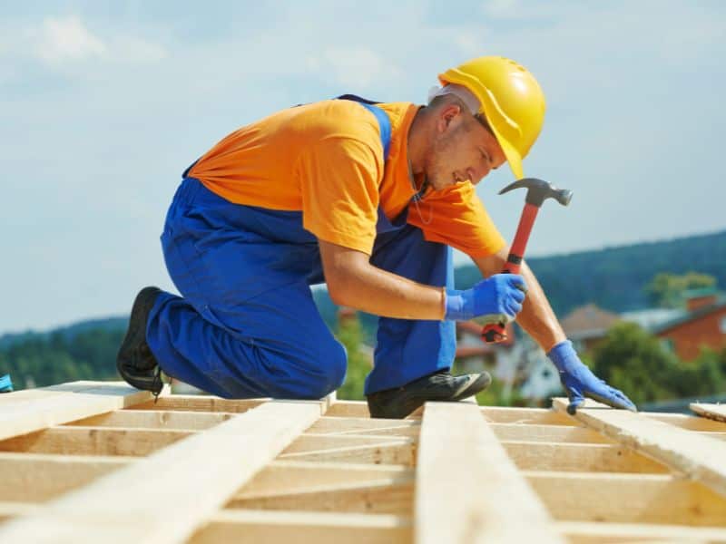 A worker wearing blue dungarees, orange T-shirt and a yellow hard hat nails wooden planks to a flat roof.