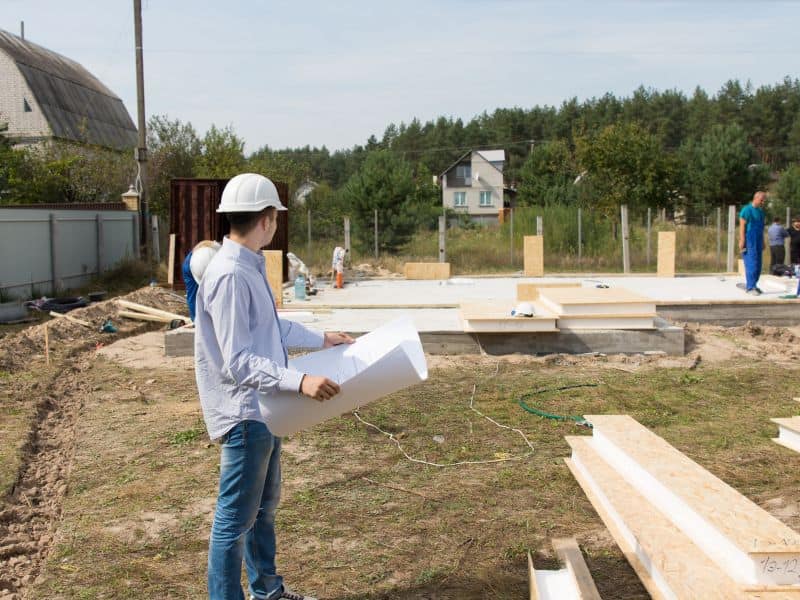 A man wearing a white hard hat holding a set of plans inspects the foundations of a new house.