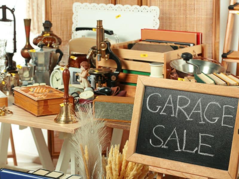 A photograph of items set out for sale at a garage sale. A sign made from a blackboard has "Garage Sale"  written in chalk. Some items for sale include a microscope, a hand bell, a wooden crate full of old records, and various ornaments.