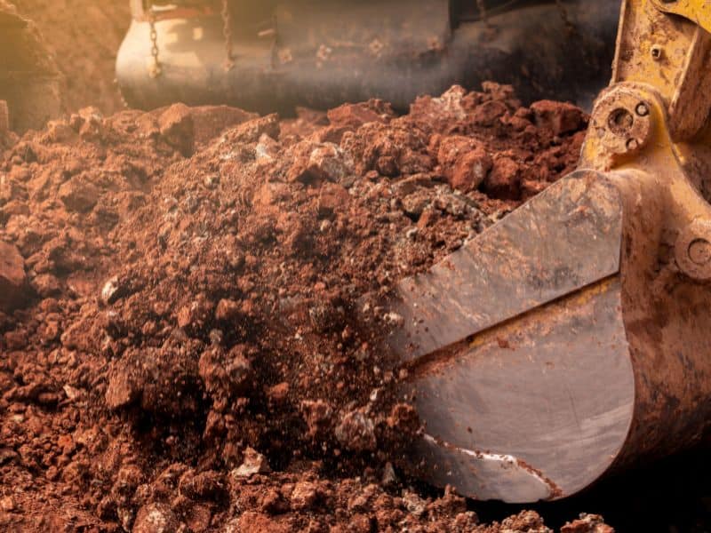 An excavator bucket digging reddish loamy subsoil.