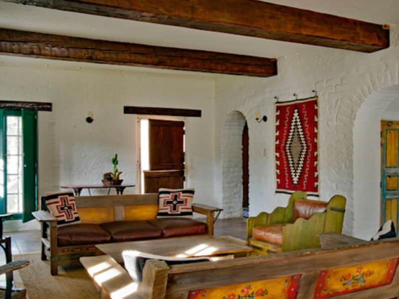 A living room in an adobe house with white walls, exposed wooden beams in the ceiling, textile wall hangings and colorful furniture.